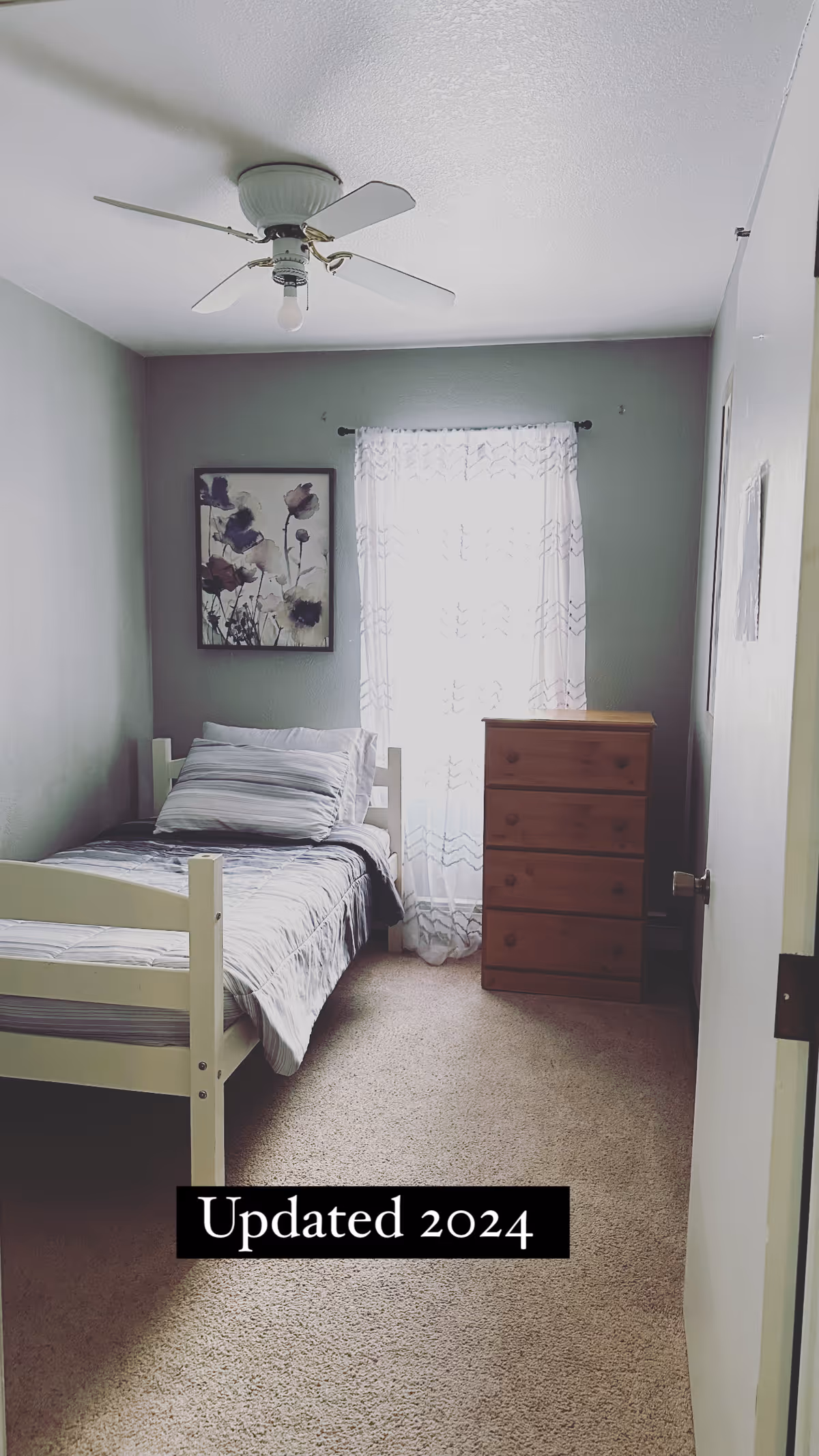 Small single bedroom with a bed, striped bedding, wooden dresser, floral wall art, sheer window curtain, and ceiling fan.