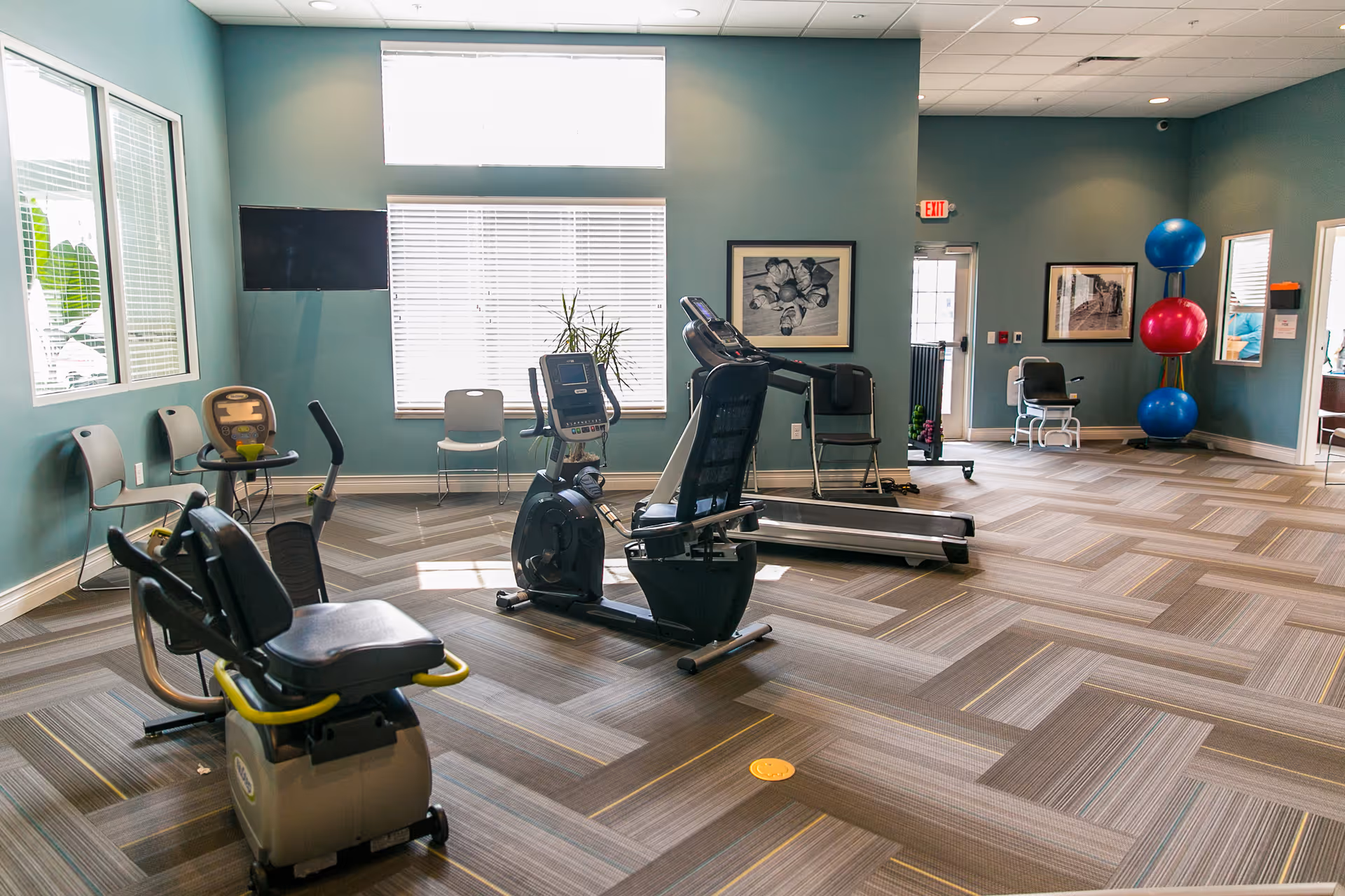 A senior fitness room with exercise equipment including a recumbent bike, treadmill, and stationary bike. The room has large windows with blinds, teal walls, framed artwork, chairs along the walls, and a rack holding large exercise balls in blue and pink. The floor has a patterned carpet and there is an exit door in the background.