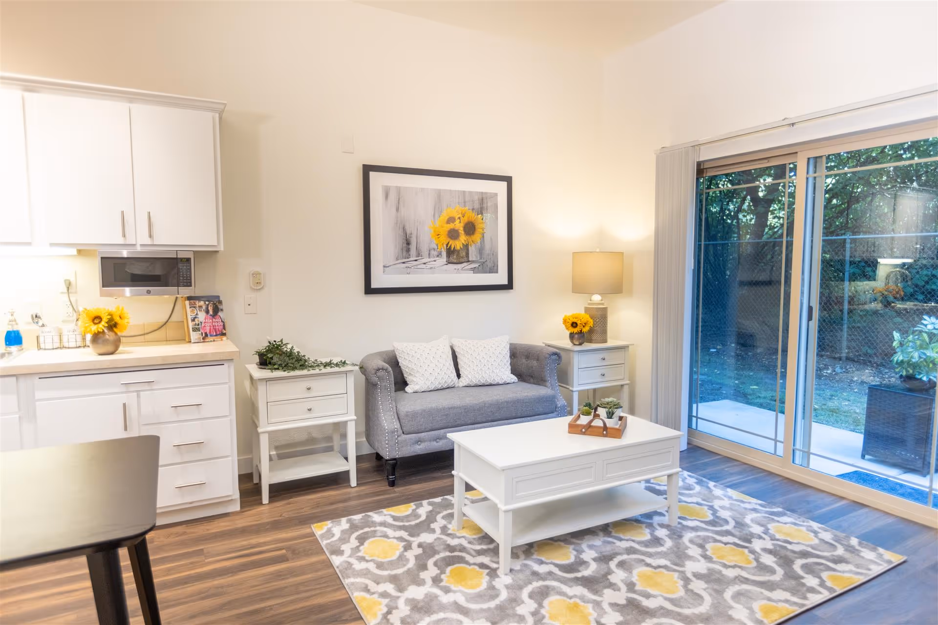 A cozy living room area with a gray loveseat adorned with two white pillows, a white coffee table with small plants on top, and a patterned rug in gray, white, and yellow. To the left, there is a kitchen counter with white cabinets, a microwave, and a vase with sunflowers. On the right side, there is a sliding glass door leading to an outdoor area with trees and a fence. A side table with a lamp and another vase of sunflowers is next to the loveseat, and a framed picture of sunflowers hangs on the wall above.