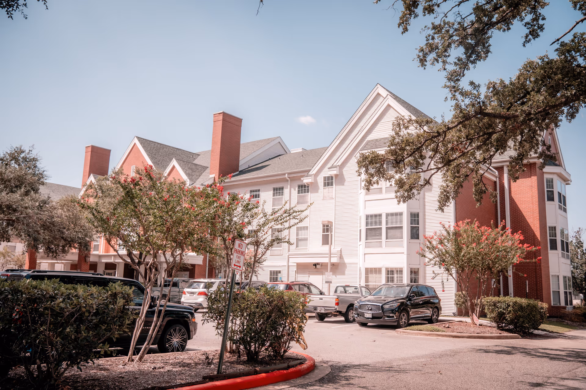 Exterior view of a senior living facility building with white siding and red brick accents, multiple windows, and a pitched roof. Several cars are parked in front of the building, surrounded by trees and shrubs under a clear blue sky.
