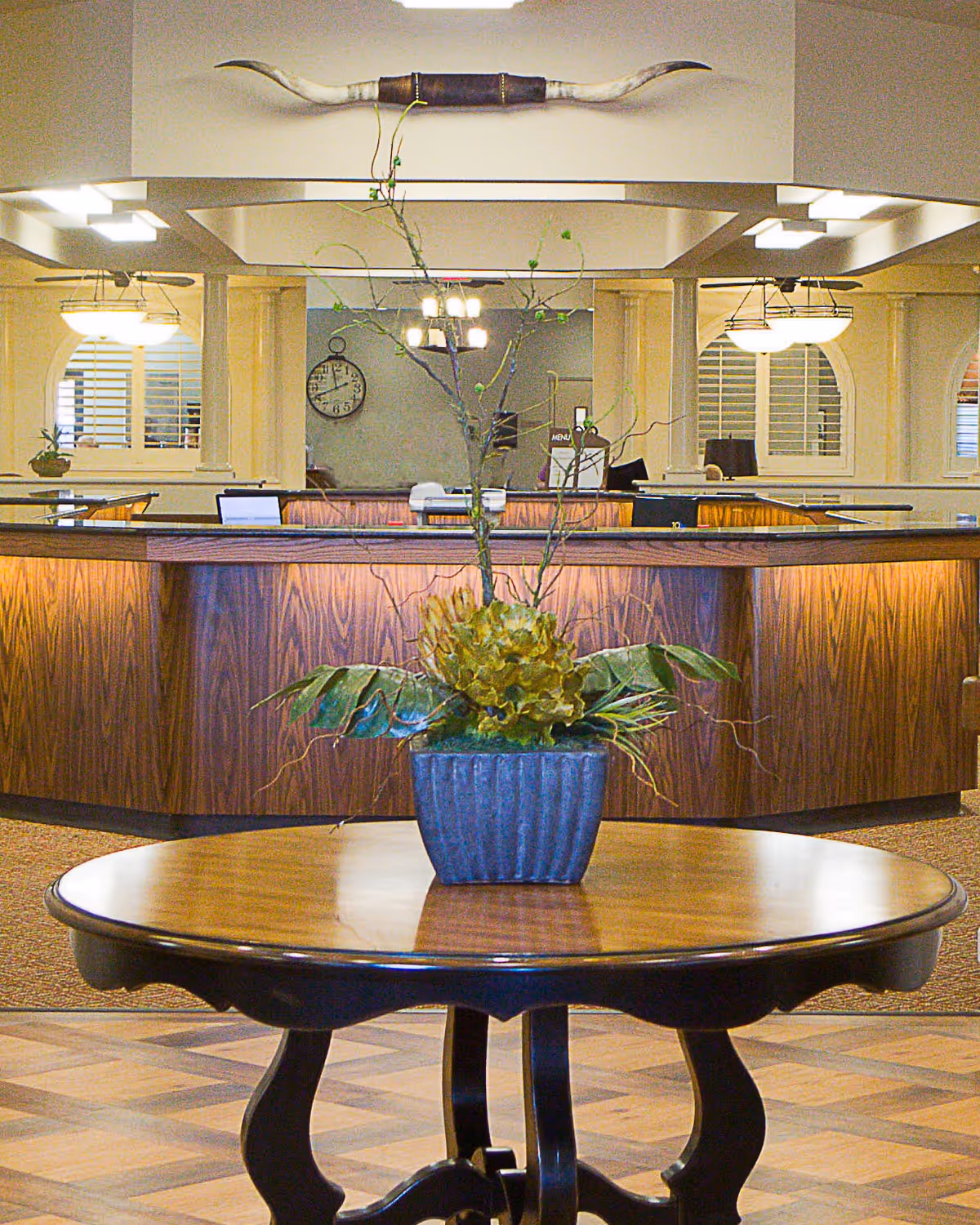 Interior view of a reception area with a round wooden table in the foreground holding a potted plant with green leaves and branches. Behind the table is a curved wooden reception desk with office equipment and a clock on the wall. The ceiling has decorative lighting and a mounted longhorn decor piece above the desk.