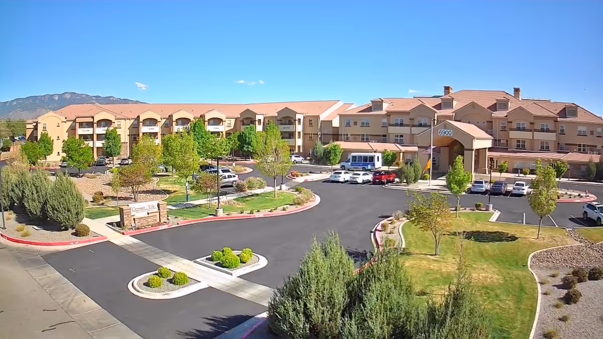 Exterior view of Cactus Valley Retirement Resort, a large multi-story building with beige walls and red-tiled roofs, surrounded by landscaped greenery, trees, and a parking lot with several cars. Mountains are visible in the background under a clear blue sky.