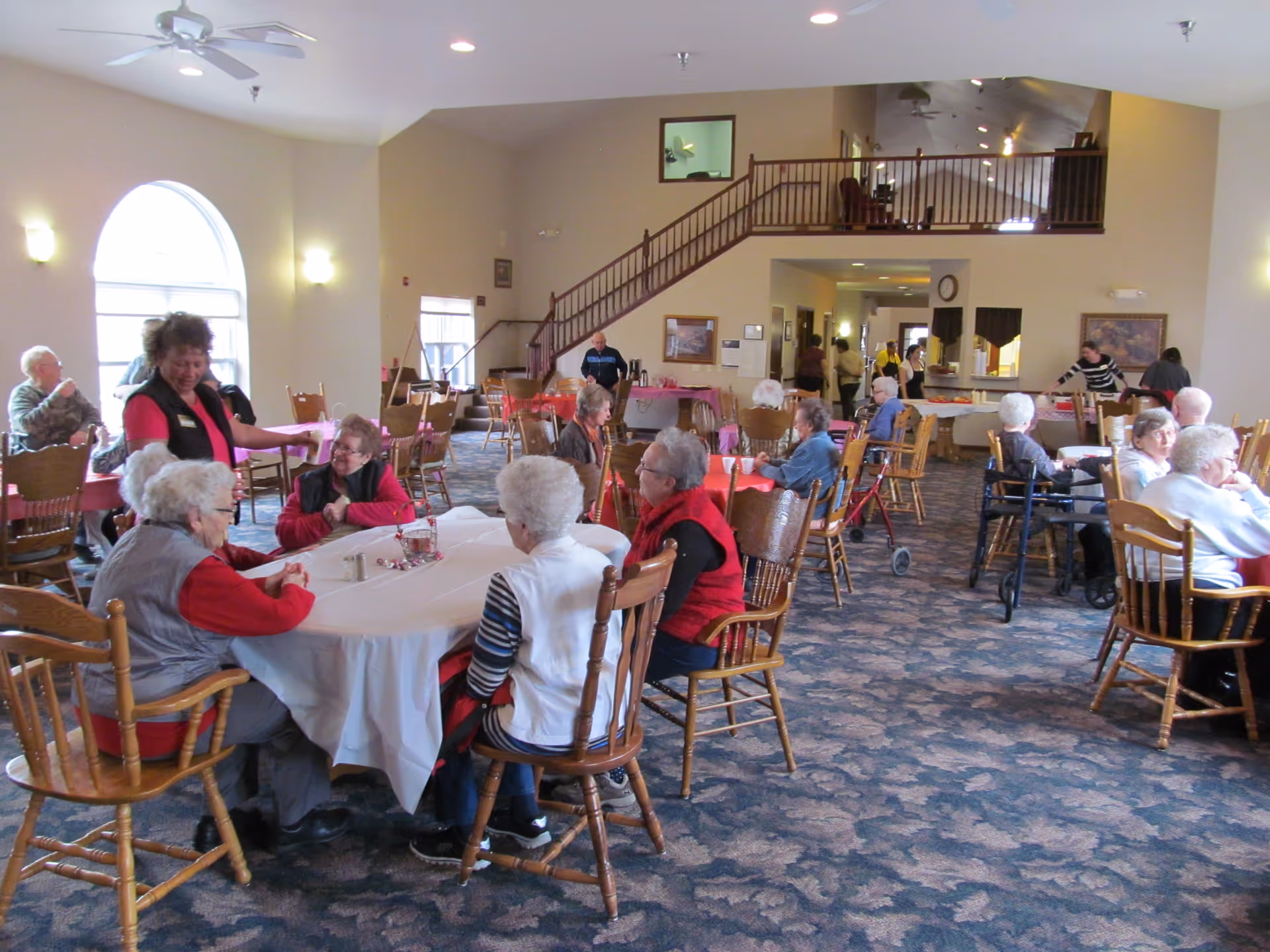 A spacious dining room in an assisted living facility with several elderly residents seated at tables covered with white and red tablecloths. A staff member is interacting with some of the residents. The room has large arched windows, a staircase leading to an upper level, and a carpeted floor with a floral pattern.