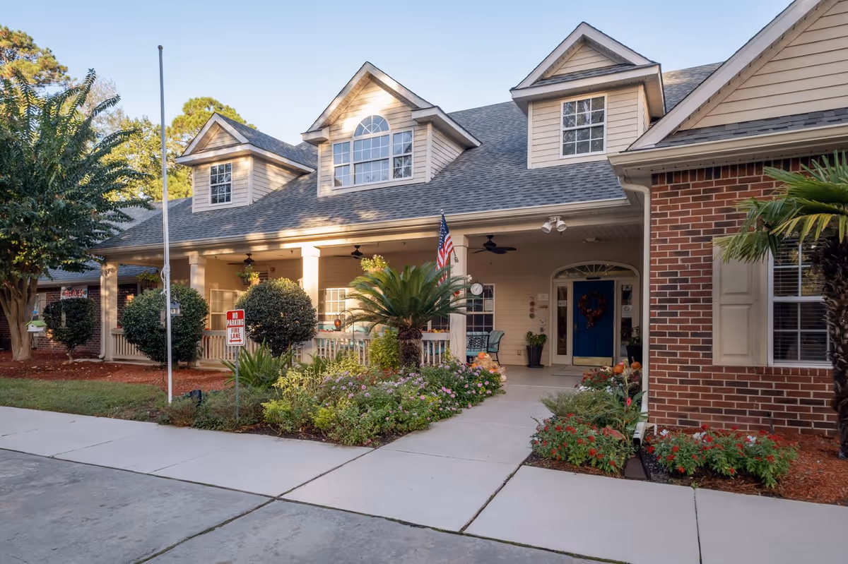 Exterior view of a residential-style assisted living facility with a covered porch, garden with various plants and flowers, and a blue front door. The building has a combination of brick and siding with dormer windows on the roof. An American flag is displayed near the entrance.