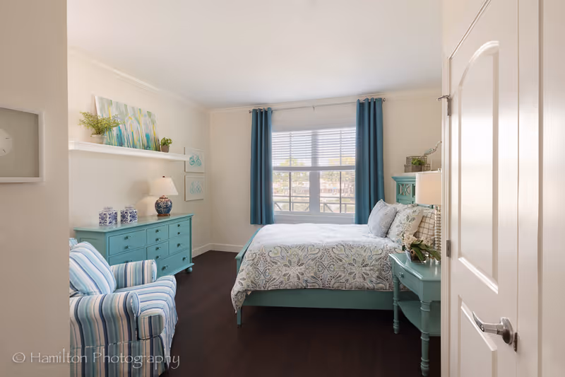 A bright and cozy bedroom with a large window covered by blue curtains. The room features a bed with patterned bedding, a matching blue nightstand with a lamp, a blue dresser with decorative items, and a striped armchair. The walls are light-colored, and there are framed artworks and plants adding a touch of greenery.