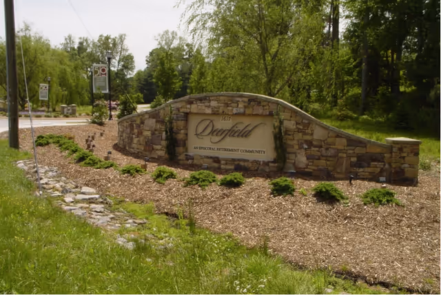 Stone entrance sign for Deerfield Episcopal Retirement Community surrounded by mulch and small green shrubs, with trees and a road in the background.