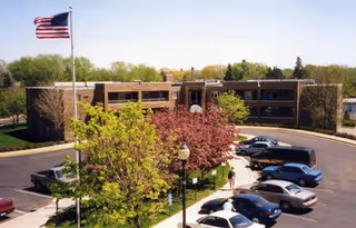 Exterior view of a two-story senior living facility building with a parking lot in front. Several cars are parked, and there are trees with green and pink foliage near the entrance. An American flag is flying on a flagpole to the left side of the image.