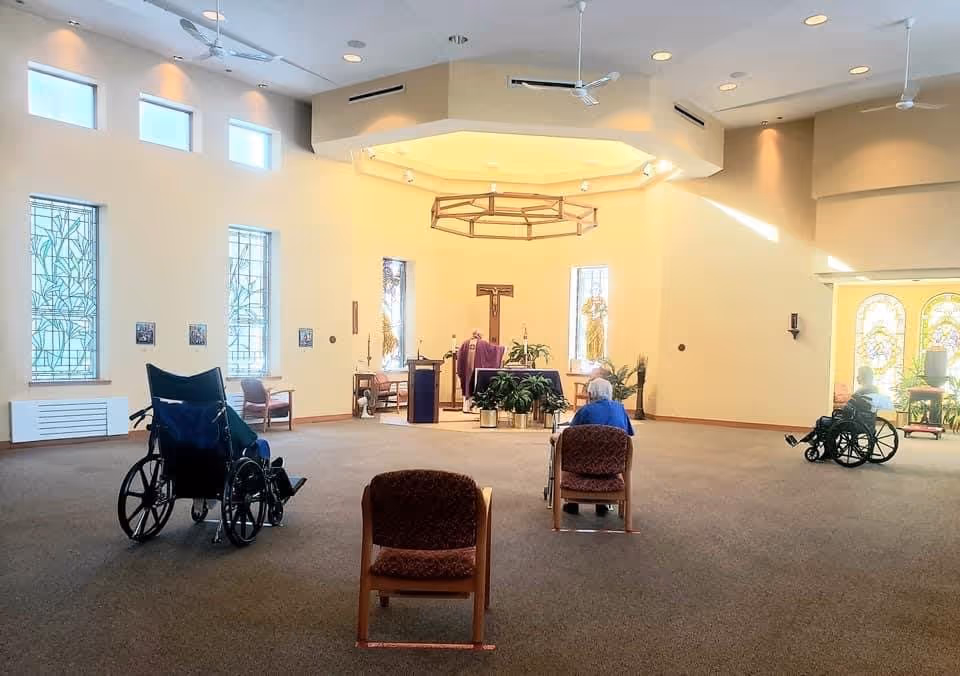 Interior of a chapel or worship room with stained glass windows and a cross at the front. There are a few elderly individuals seated, some in wheelchairs and some in chairs, facing the altar where a person is standing. The room is spacious with high ceilings and soft lighting.