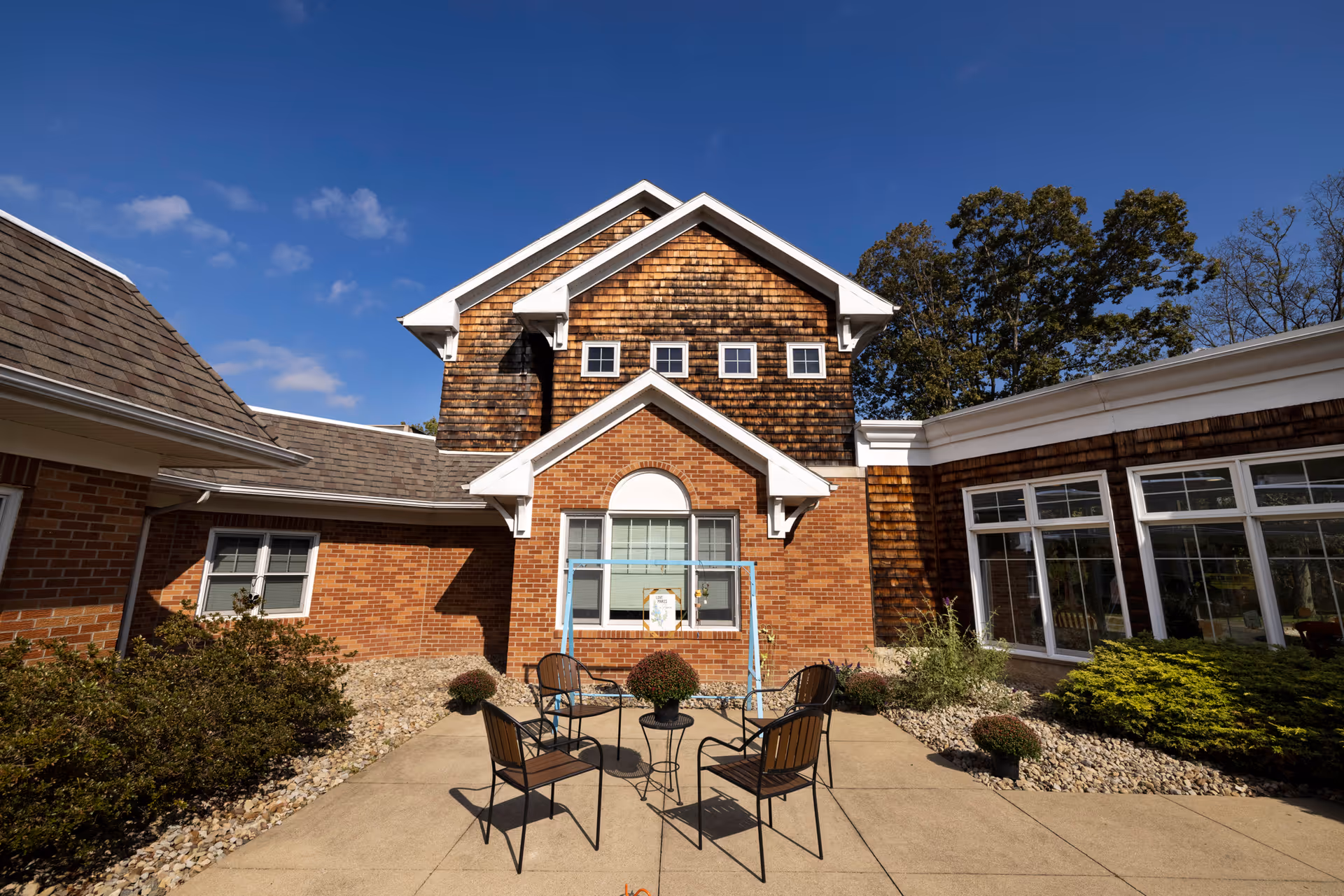 Outdoor courtyard area of Carroll Senior Care Facility with four metal chairs arranged around a small round table on a concrete patio. The building features a combination of brick and wood siding with multiple windows and a clear blue sky above.