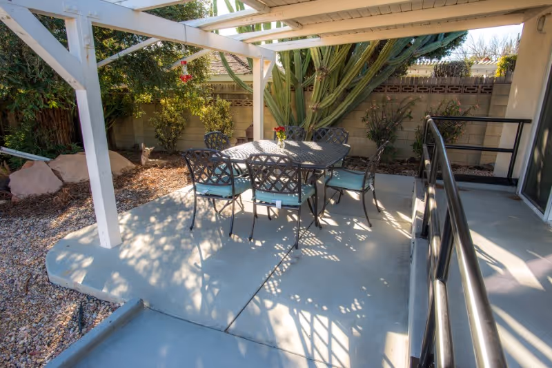 Outdoor patio area with a metal table and six chairs with cushions under a white pergola. The patio is surrounded by desert landscaping with rocks, plants, and a large cactus against a block wall fence. There is a black metal handrail along a concrete ramp leading to a sliding glass door.