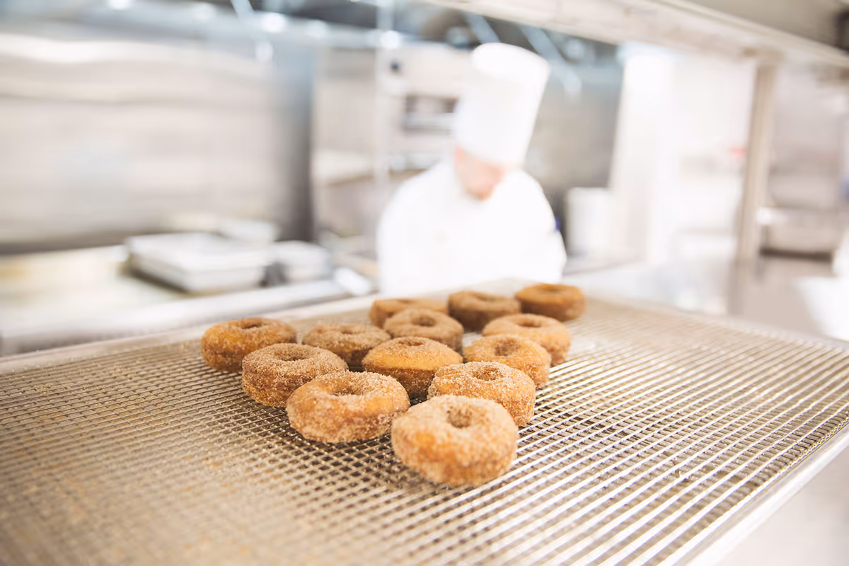 A tray of freshly made cinnamon sugar donuts cooling on a wire rack in a commercial kitchen with a chef in white uniform and hat blurred in the background.