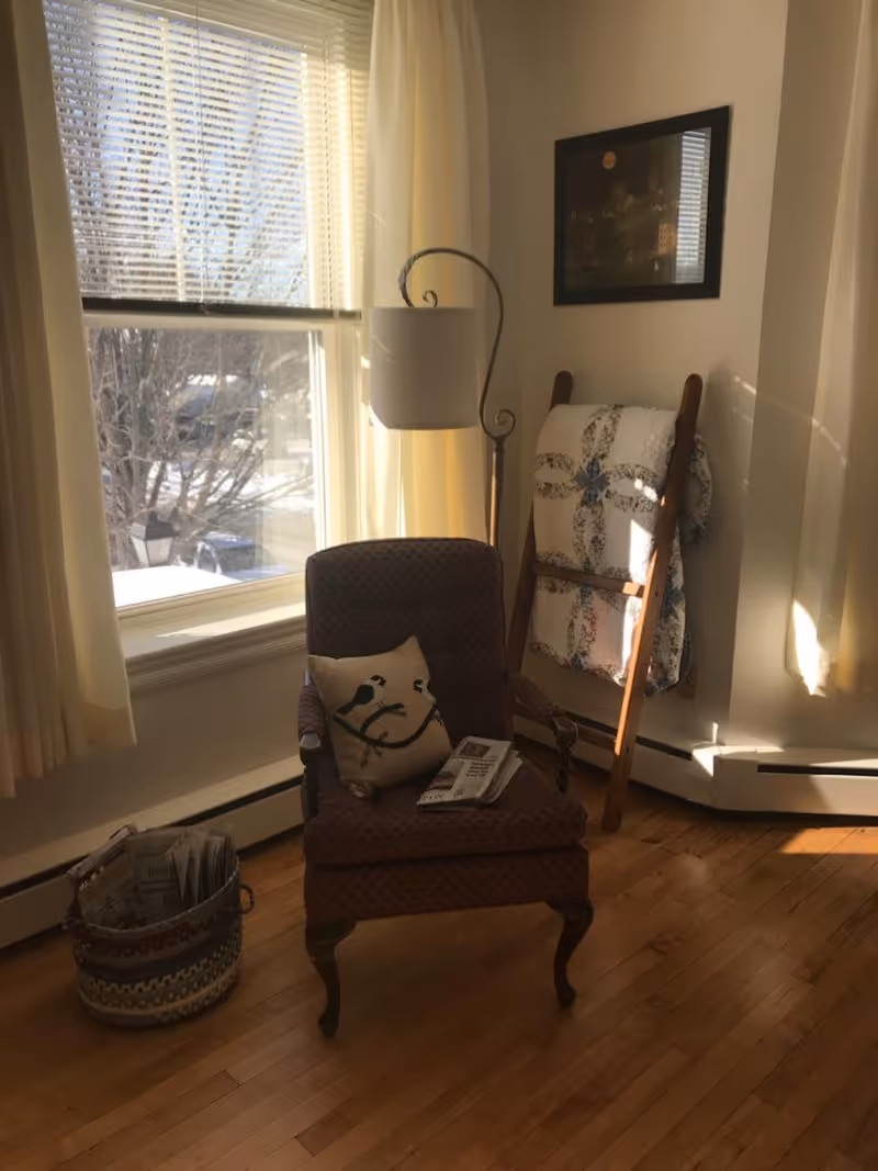 A cozy corner of a room with a patterned armchair featuring a bird-themed pillow and a folded newspaper on the seat. Next to the chair is a basket filled with newspapers. Behind the chair is a tall floor lamp and a wooden quilt rack holding a patterned quilt. Sunlight streams through a large window with blinds and light curtains, illuminating the hardwood floor.
