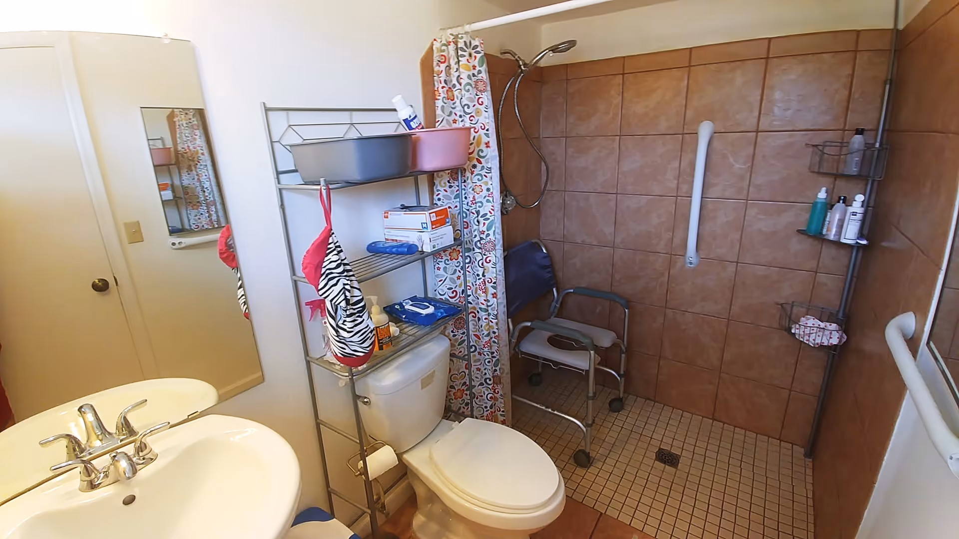 A bathroom with a white sink and faucet, a toilet with a metal shelving unit above it holding various items, and a walk-in shower area with brown tiled walls, a shower chair, grab bars, and a colorful shower curtain.