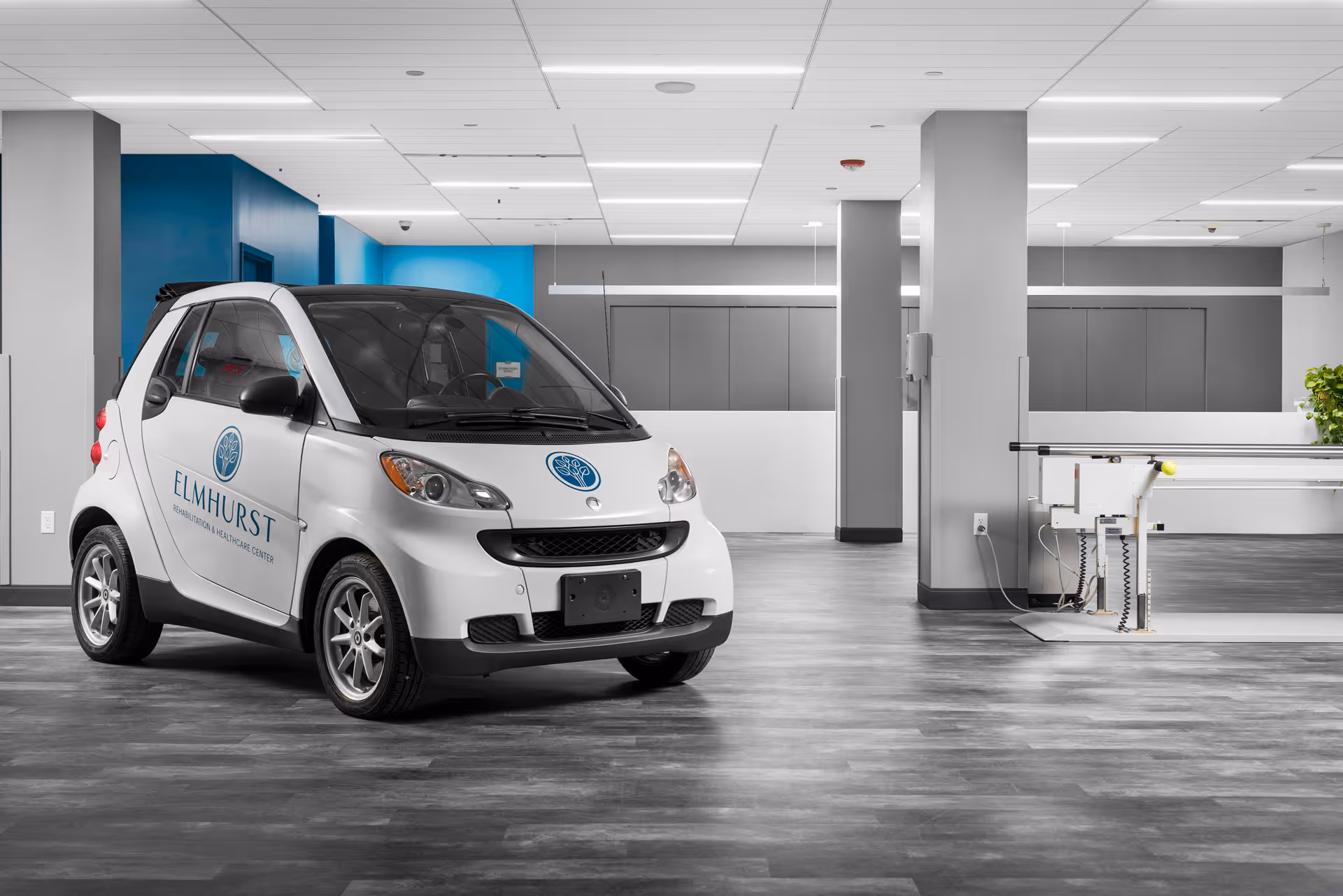 A small white Smart car with Elmhurst Rehabilitation & Healthcare Center logos on the door and hood is parked inside a spacious, modern room with gray flooring, gray and blue walls, and white ceiling panels. The room also contains parallel bars used for physical therapy on the right side.