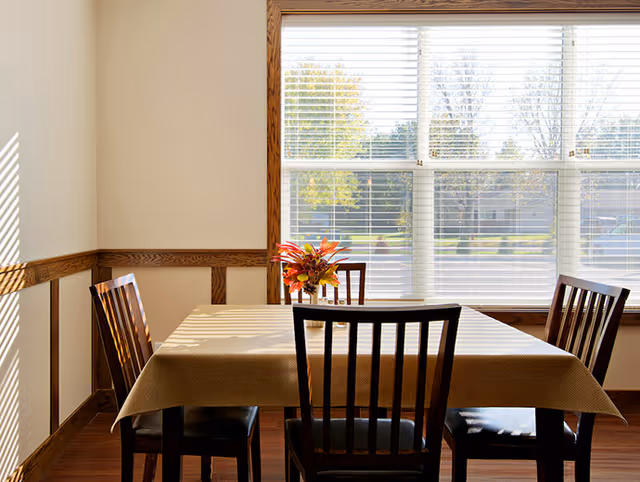 Sunlit dining table with four wooden chairs and a small vase of flowers in front of a large window.