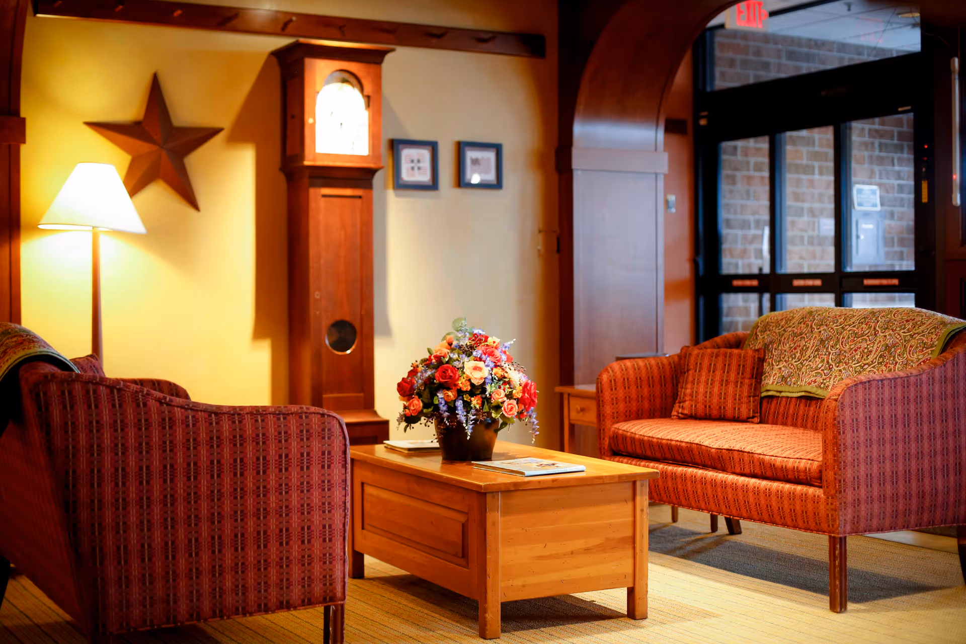 A cozy living room area with two red patterned armchairs facing a wooden coffee table adorned with a colorful flower arrangement. A tall wooden grandfather clock stands against a yellow wall, accompanied by a floor lamp and a decorative star. The background shows a glass door entrance with brick walls outside.