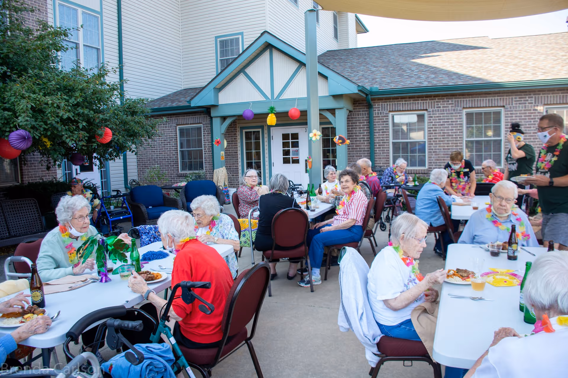 A group of elderly people sitting at tables outdoors, enjoying a meal together. The setting is a courtyard of a senior living facility with brick walls and windows in the background. Some people are wearing colorful leis and masks, and there are festive decorations hanging around.