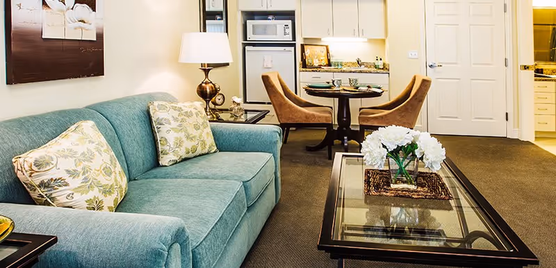 Cozy living area with a teal sofa, glass coffee table topped with white flowers, and a small dining table and kitchenette in the background.