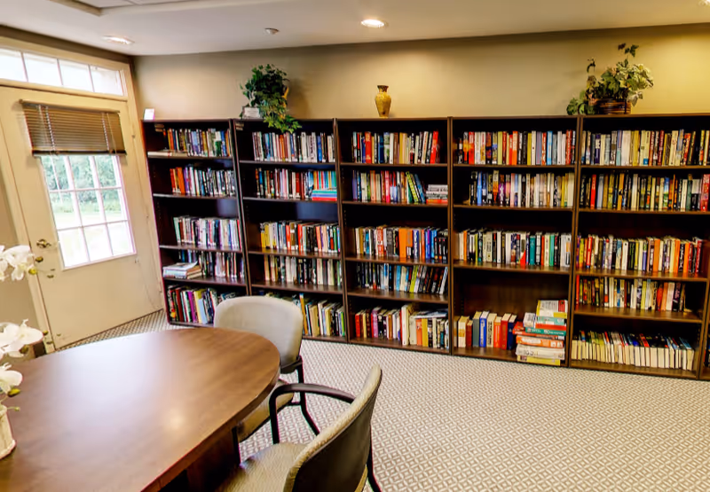A cozy reading room with wall-length bookshelves, a round table and chairs near a door and carpeted floor.