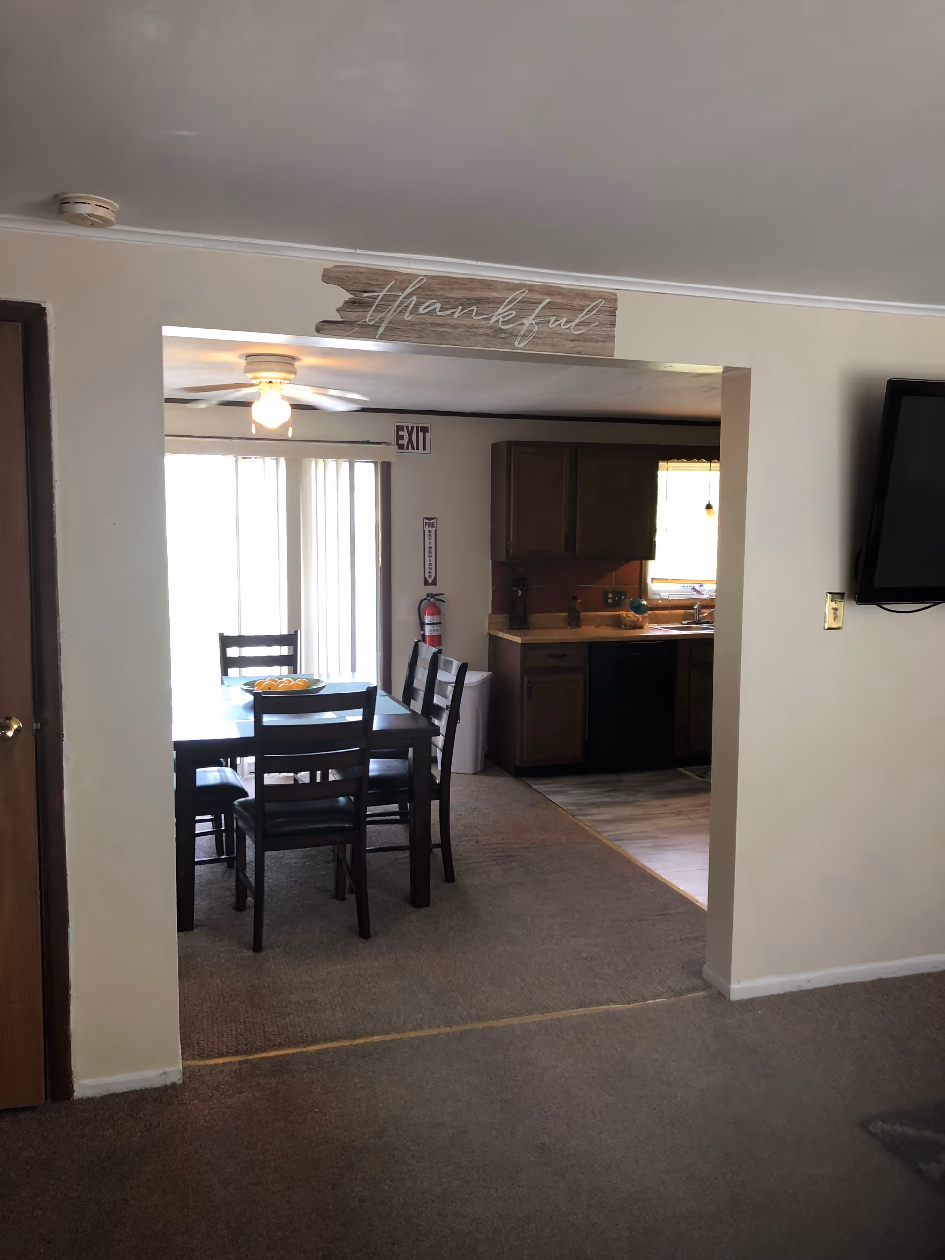 View of a dining area with a dark wooden table and four chairs, adjacent to a kitchen with wooden cabinets and a window. A ceiling fan with a light is above the dining table, and a sliding glass door with vertical blinds is in the background. A fire extinguisher is mounted on the wall near the exit sign. The walls are light-colored, and a wooden sign with the word 'thankful' is above the doorway between the dining area and the kitchen.