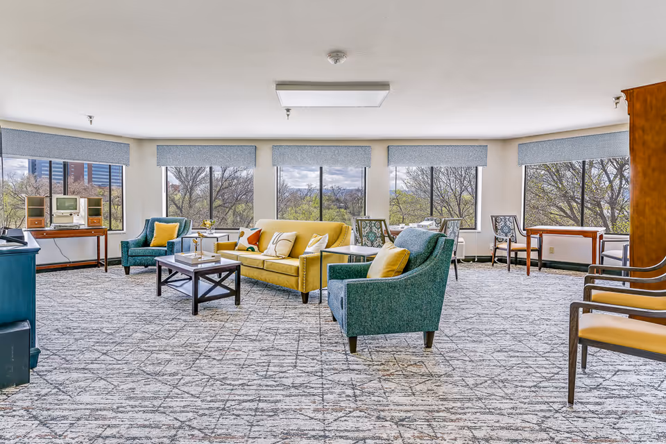A spacious senior living common area with large windows showing outdoor trees and buildings. The room features a yellow sofa with colorful pillows, two teal armchairs with yellow pillows, a wooden coffee table, and several wooden tables and chairs along the windows. The carpet has a geometric pattern, and the walls are light-colored with blue window valances.