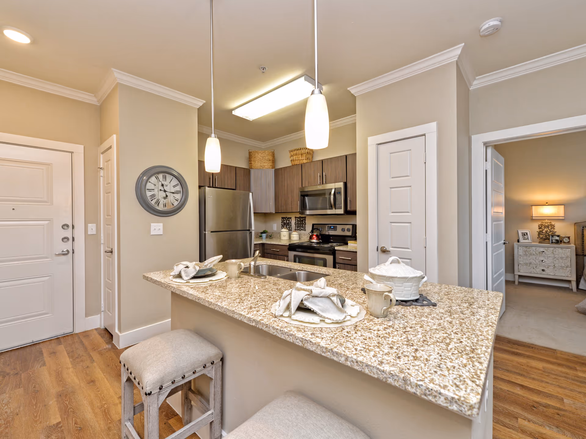 Interior view of a modern kitchen with a granite countertop island set with plates, mugs, and napkins. The kitchen features stainless steel appliances including a refrigerator, stove, and microwave. Two pendant lights hang above the island. Adjacent to the kitchen is a doorway leading to a bedroom with a nightstand and lamp visible.