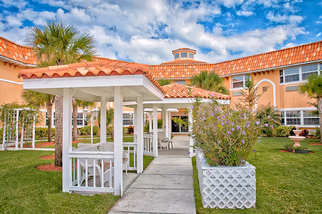 Courtyard of Heron House Assisted Living with white covered walkways and gazebos, a tile-roofed building, palm trees, and landscaped lawn under a blue sky.