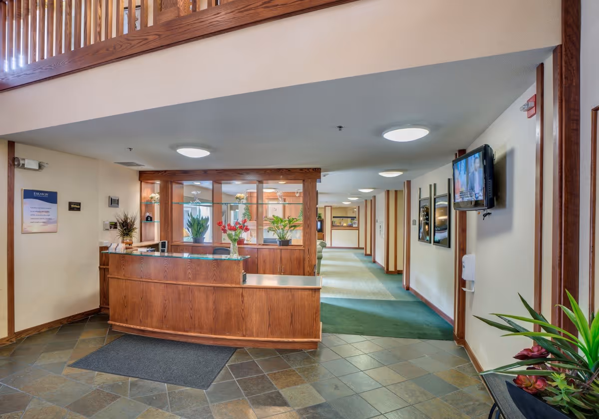 Reception area with a wooden front desk, plants, and a hallway leading to other rooms. The floor is tiled near the desk and carpeted in the hallway. A TV is mounted on the wall to the right, and there are framed pictures and a hand sanitizer dispenser on the wall.