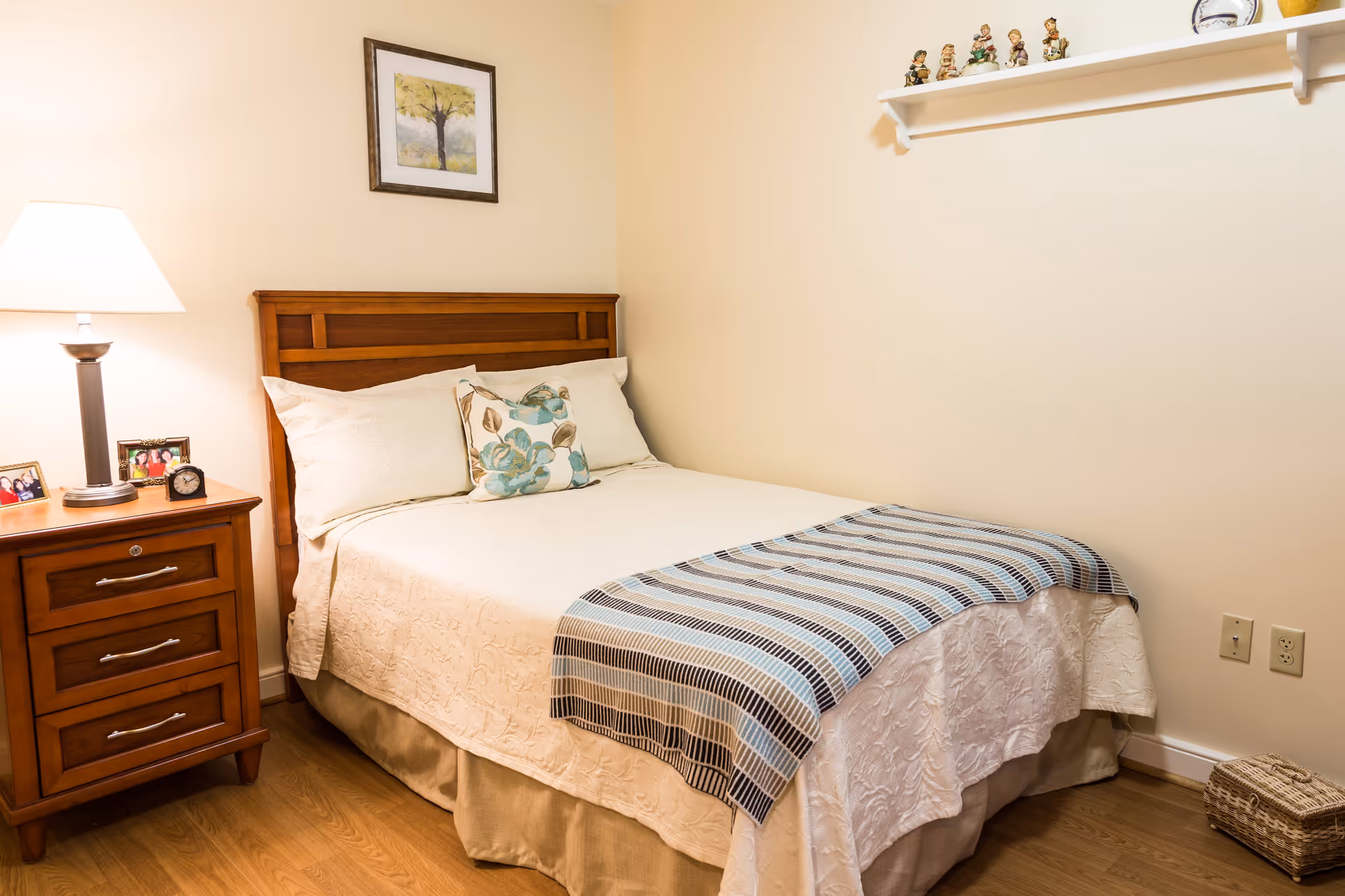A cozy bedroom with a wooden bed frame, white bedding, and a striped blue and brown throw blanket. There is a wooden nightstand with a lamp, framed photos, and a small clock. A framed picture hangs on the wall above the bed, and a shelf with small figurines is mounted on the adjacent wall. The floor is wooden, and a woven basket is placed near the wall.