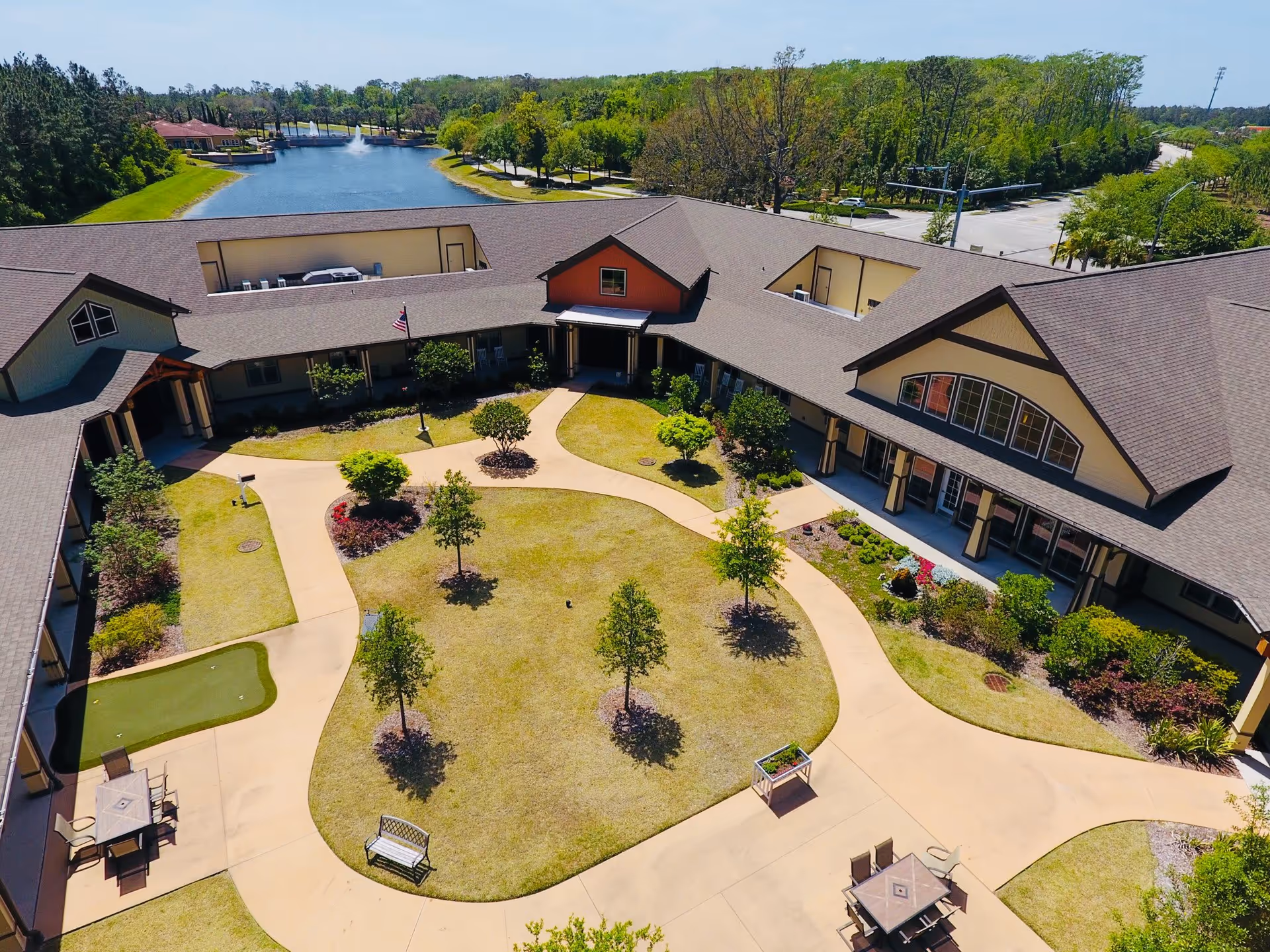 Aerial view of a senior living facility courtyard with a well-maintained lawn, small trees, walking paths, outdoor seating areas, and a putting green. The building surrounds the courtyard in a U-shape, and there is a pond with a fountain visible in the background along with trees and a road.