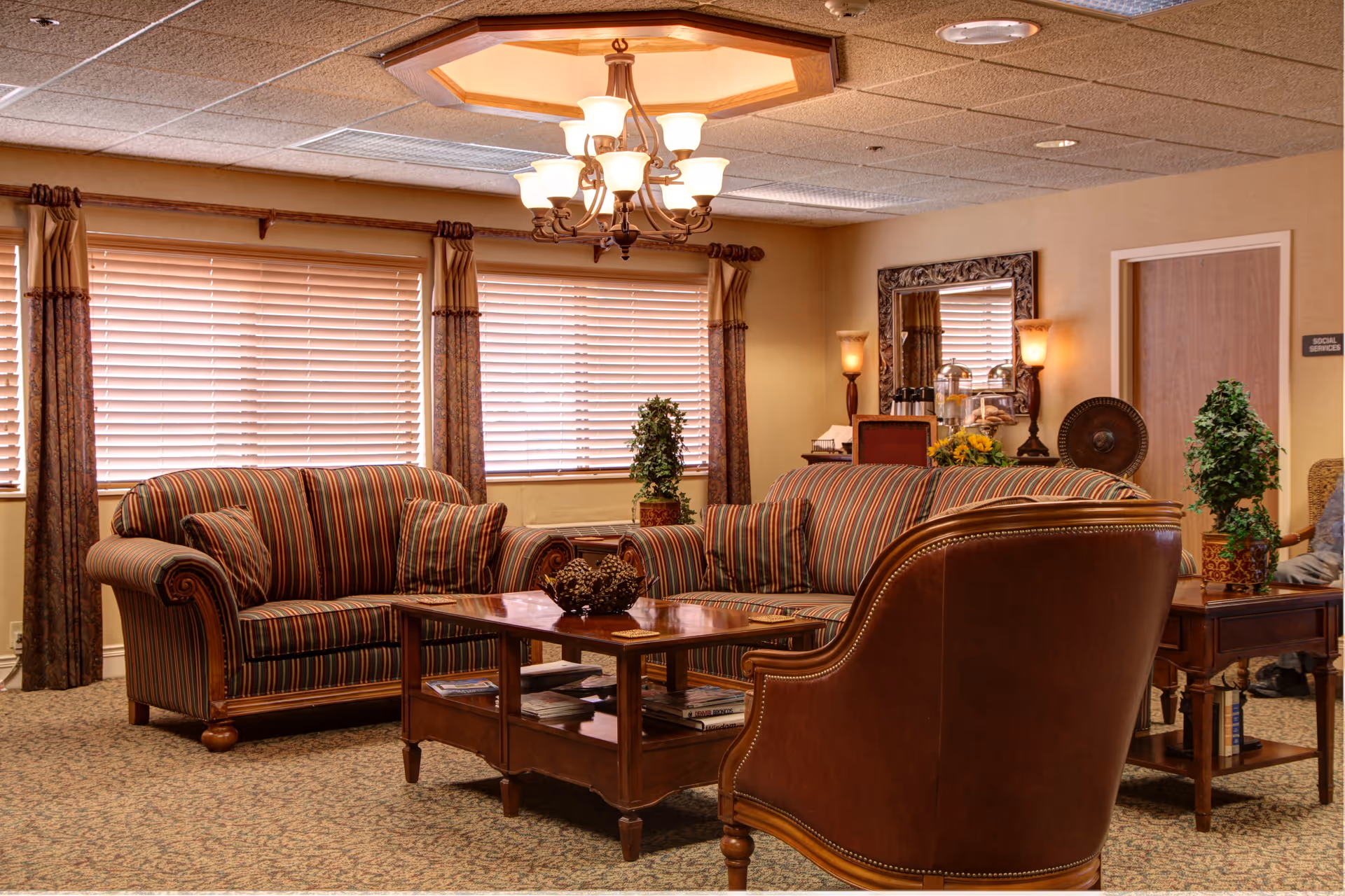 A cozy living room area with striped upholstered sofas and a leather armchair arranged around a wooden coffee table. The room features large windows with blinds and curtains, a decorative chandelier, a large mirror on the wall, potted plants, and warm lighting from wall sconces.