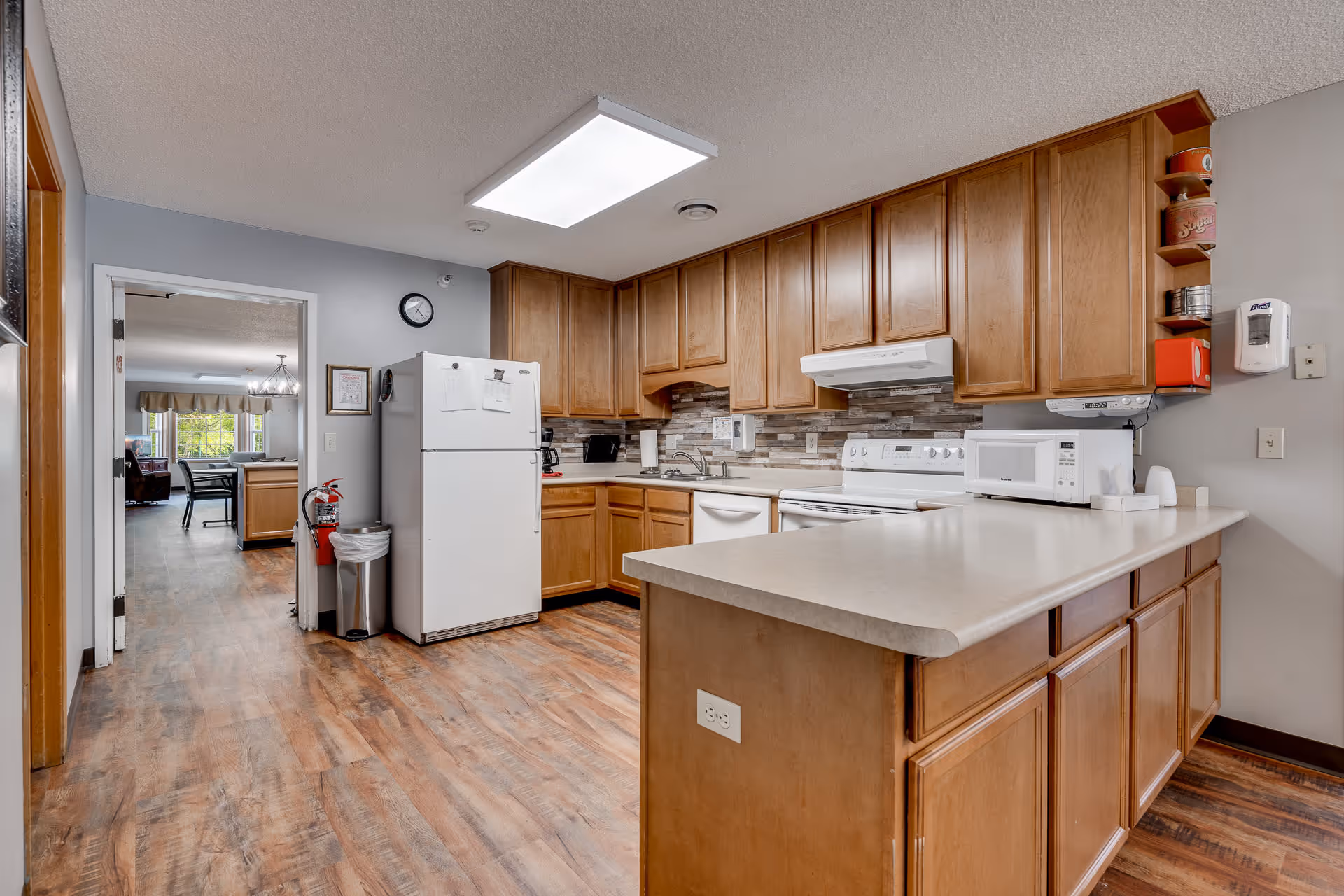 A spacious kitchen with wooden cabinets, a white refrigerator, stove, microwave, and dishwasher. The kitchen has a light-colored countertop island and wood-patterned flooring. A doorway leads to a dining area with a table and chairs, and a large window with curtains.