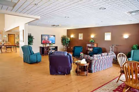 A senior living lounge with teal chairs, a patterned sofa, coffee tables, and a TV.