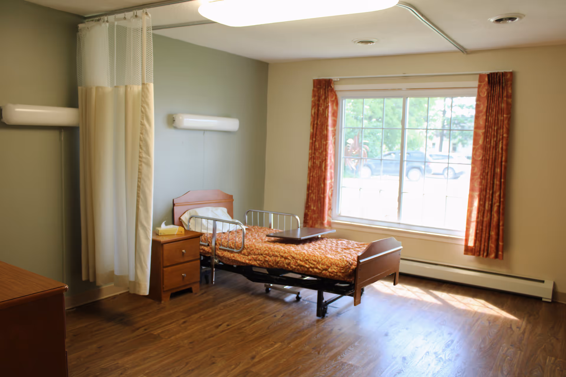 A senior care patient room with a single adjustable bed, bedside table, privacy curtain and a large window with curtains.