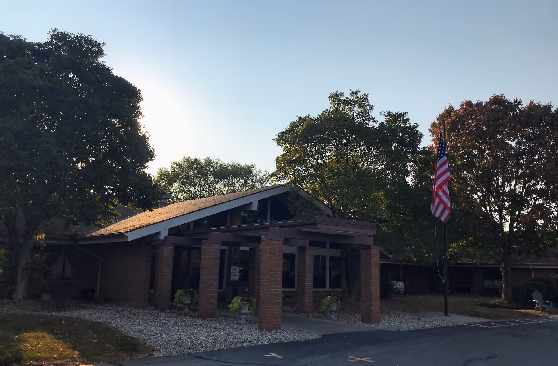 Exterior view of a single-story brick building with a sloped roof, surrounded by trees and a paved driveway. An American flag is displayed on a flagpole near the entrance, which has a covered porch supported by brick columns.