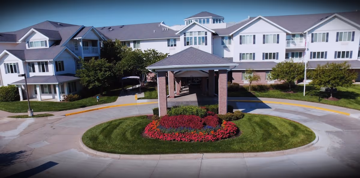 Front exterior view of a senior living facility with a circular driveway and a landscaped island featuring red and green plants. The building is white with multiple windows and a covered entrance supported by brick columns.