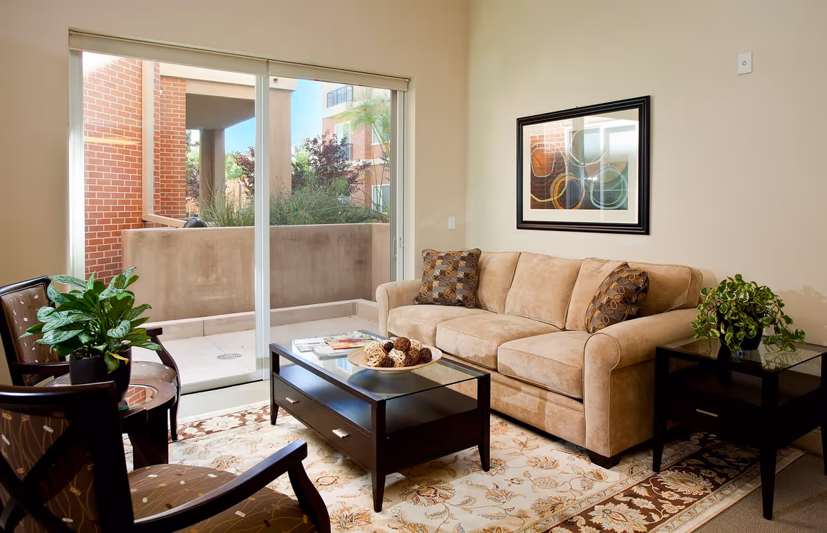 A cozy living room with a beige sofa adorned with two patterned cushions, a glass-top coffee table with decorative balls, two wooden chairs with patterned cushions, a side table with a green potted plant, a large window with sliding glass doors leading to a small outdoor patio, and a framed abstract artwork on the wall.