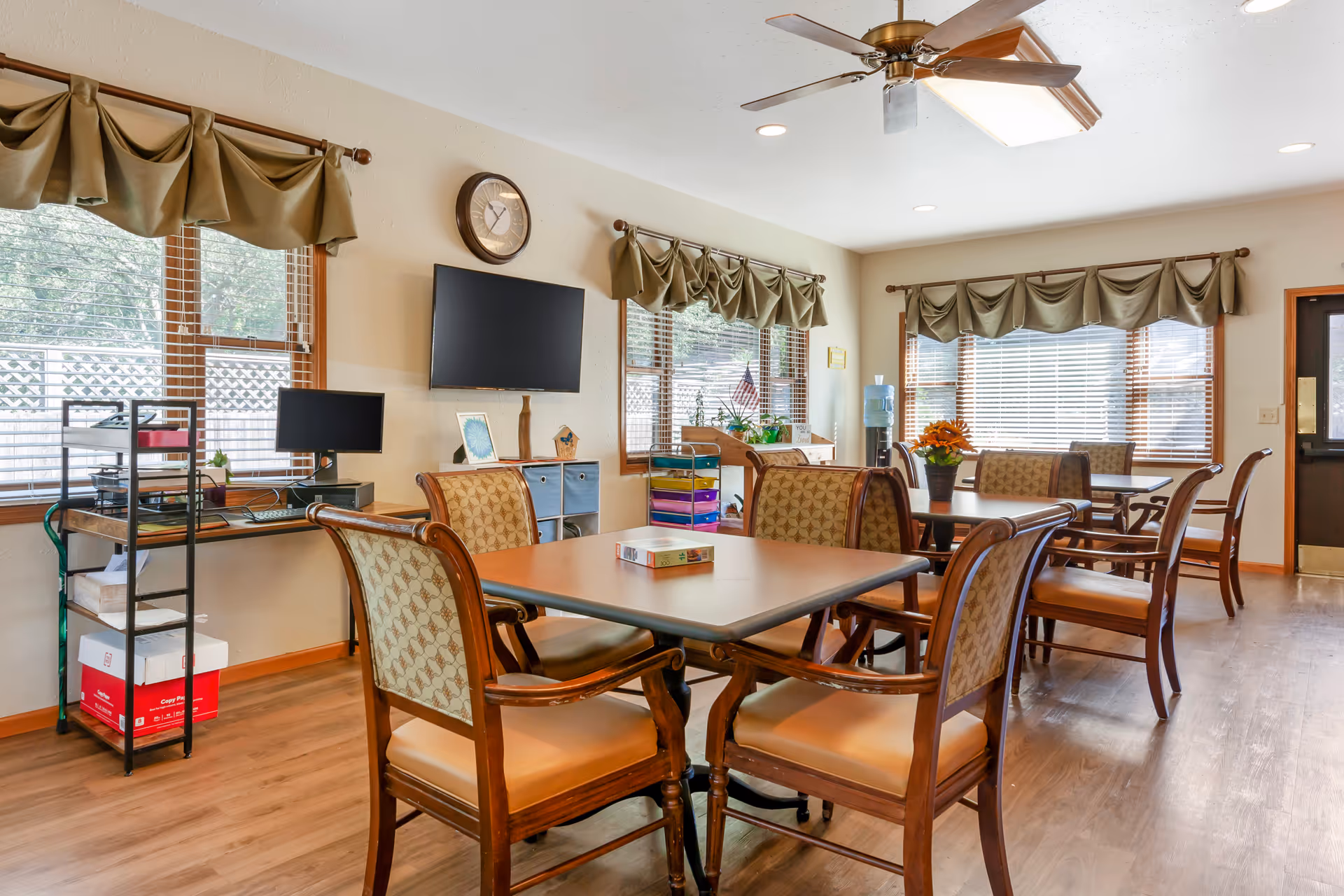 A bright common area with wooden floors and multiple tables surrounded by cushioned wooden chairs. The room has large windows with green valance curtains, a ceiling fan, a wall clock, a mounted flat-screen TV, and a water dispenser. There is a small desk with a computer and office supplies near the windows.