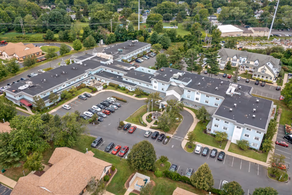 Aerial view of the Spring Hills Matawan senior living facility showing a large, multi-wing building with a black roof surrounded by parking lots, trees, and greenery. Several cars are parked in the parking areas, and the facility is situated in a suburban neighborhood with other houses and buildings nearby.