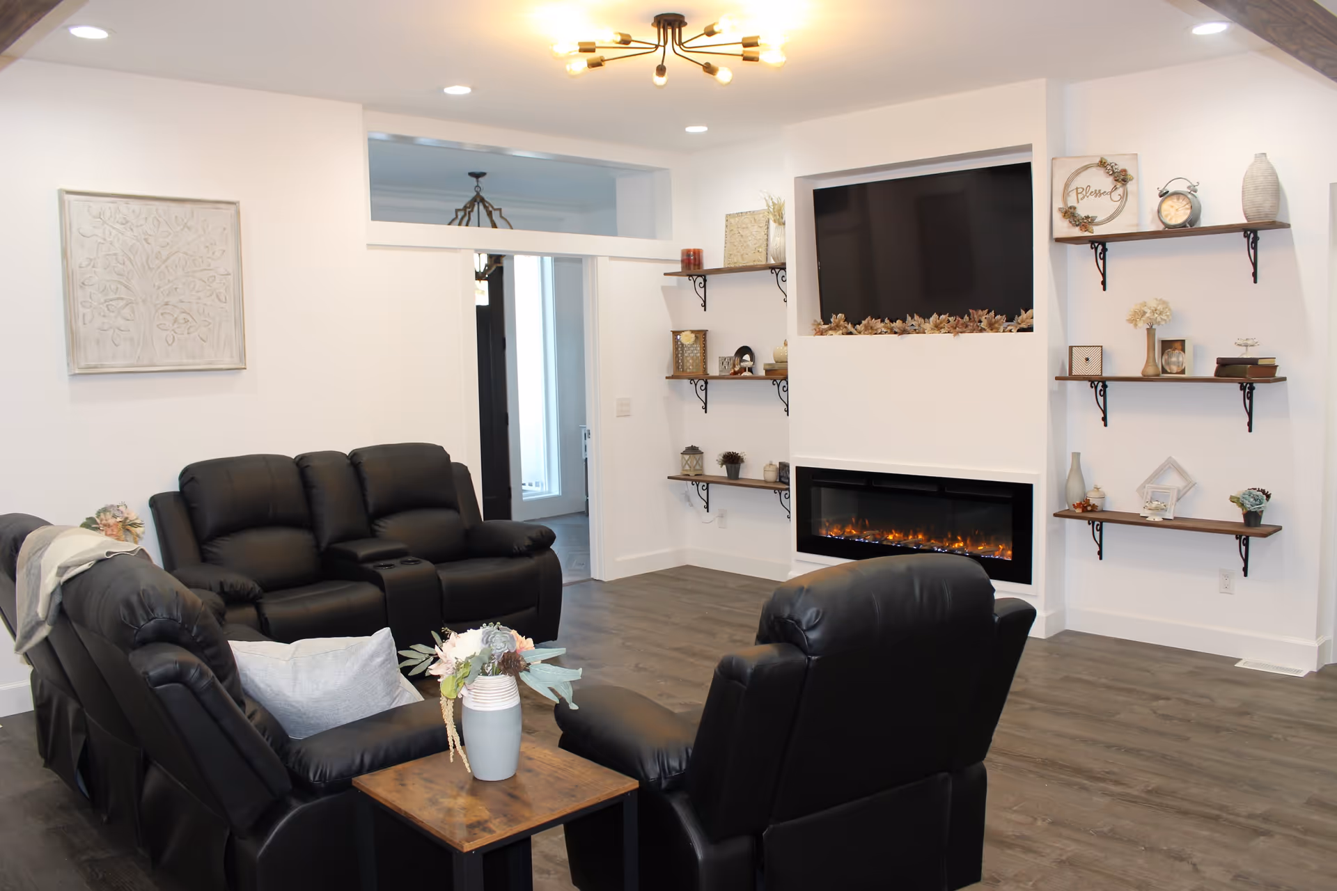 A cozy living room with black leather recliner chairs arranged around a wooden side table with a vase of flowers. The room features a modern electric fireplace beneath a mounted flat-screen TV, with decorative shelves on either side holding various ornaments. The walls are white, and the floor is a dark wood laminate.