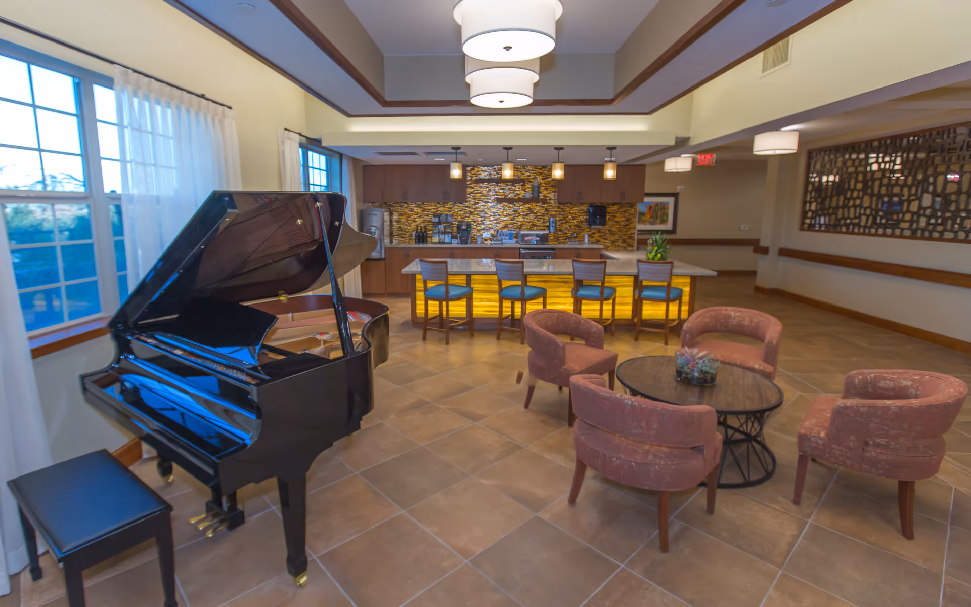 Interior view of a senior living facility lounge area featuring a black grand piano with a bench near large windows with sheer curtains, a seating area with four upholstered chairs around a round wooden table with a small plant centerpiece, and a kitchen counter with four bar stools and a decorative yellow backlit panel beneath the counter. The kitchen area has wooden cabinets and a mosaic tile backsplash.