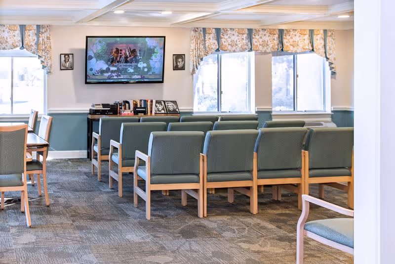 A common area with rows of green cushioned chairs facing a wall-mounted television. The room has large windows with floral valances, a carpeted floor, and a small table with books and framed photos beneath the TV.