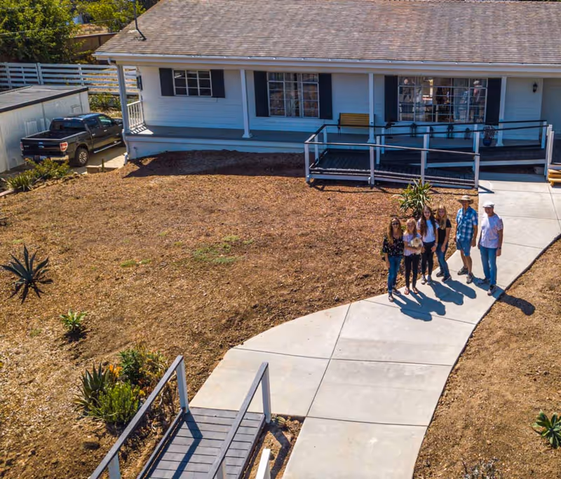 A group of six people standing on a curved concrete walkway in front of a single-story white house with a porch and ramp. The yard around the walkway is mostly bare soil with some small plants and shrubs. A black pickup truck is parked near a white shed to the left of the house.