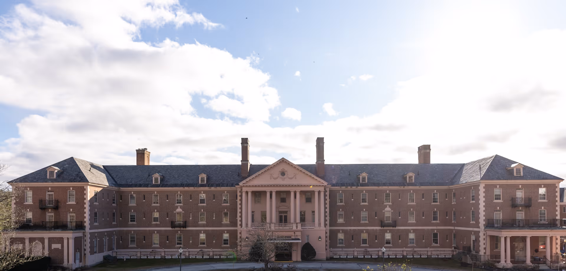 Front exterior view of a large, symmetrical, three-story brick building with a central portico supported by columns and multiple chimneys on the roof under a partly cloudy sky.