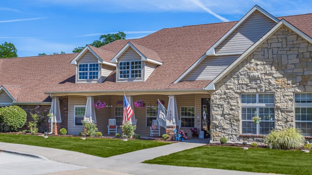 Front exterior view of a single-story senior living facility building with a brown shingled roof, beige siding, and stone facade. The entrance has a covered porch with two rocking chairs, hanging flower baskets, and two flags displayed. Well-maintained green lawn and landscaping surround the building under a clear blue sky.
