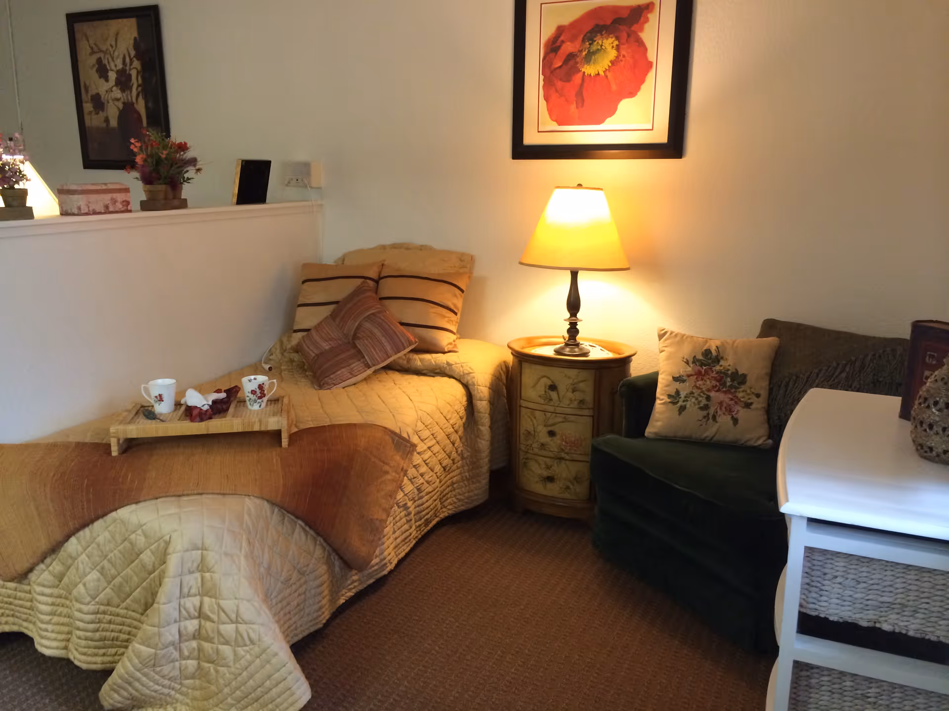 A cozy bedroom with a single bed covered in a beige quilt and several pillows. A wooden tray with two cups and a napkin is placed on the bed. Next to the bed is a small round nightstand with a lit table lamp. A green armchair with a floral pillow is beside the nightstand. On the wall above the nightstand is a framed picture of a red flower. The room has carpeted flooring and light-colored walls with another framed picture partially visible.