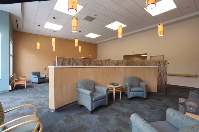 A spacious, well-lit common area with multiple armchairs and small tables arranged around a central wooden partition. The room features hanging pendant lights, large windows letting in natural light, and a patterned carpet floor.