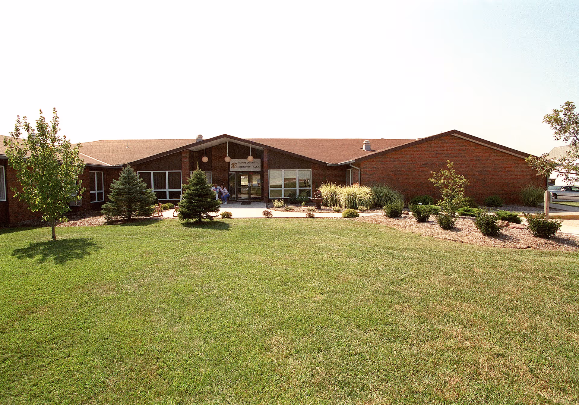 Front exterior view of a single-story brick building with a well-maintained lawn and small trees and shrubs in front. The entrance has a covered porch with hanging lights and glass doors. A few people are visible near the entrance.
