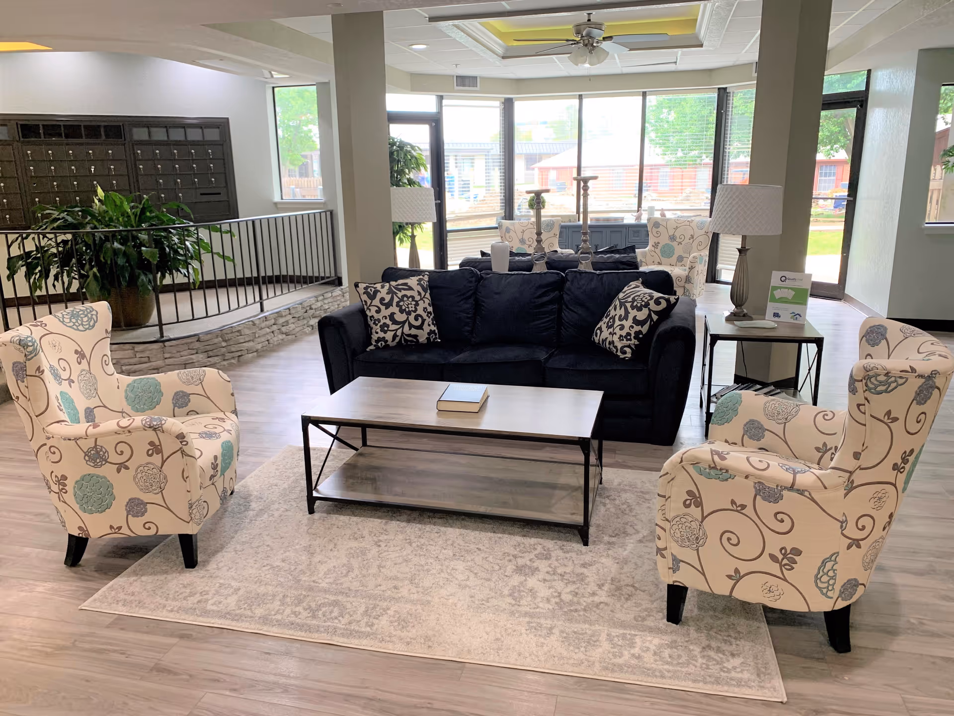 Bright communal living area with a navy sofa, patterned armchairs, a coffee table, and large windows overlooking the exterior.