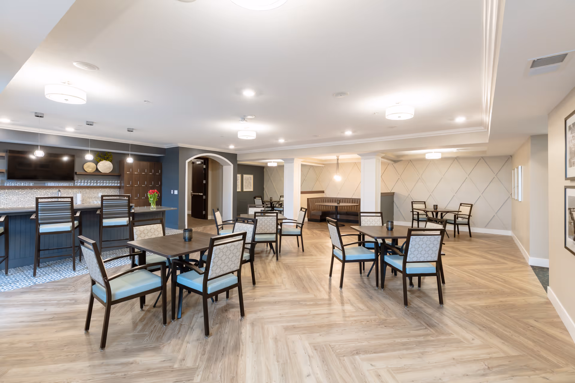 A spacious and well-lit dining area with multiple square tables and chairs arranged neatly on a light wood patterned floor. There is a bar area with high chairs and a large TV mounted on the wall. The walls are decorated with subtle patterns and framed pictures, and the ceiling has modern light fixtures.